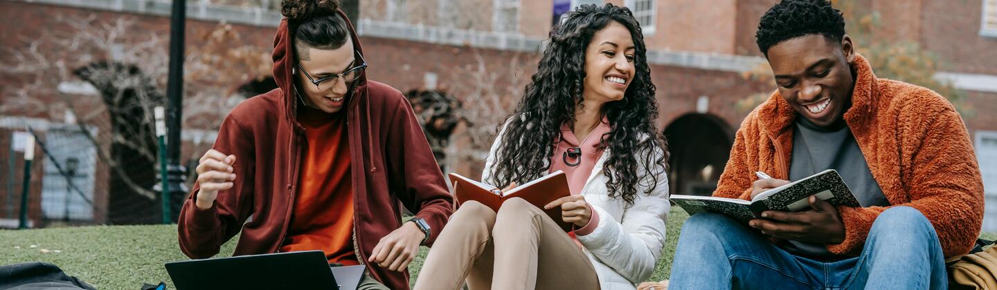 Three students laughing and studying while sitting on a grassy hill